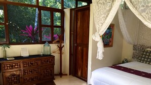 Un dormitorio con una cama, un aparador de madera, un banco con toallas, una ventana con vista a árboles y una puerta.