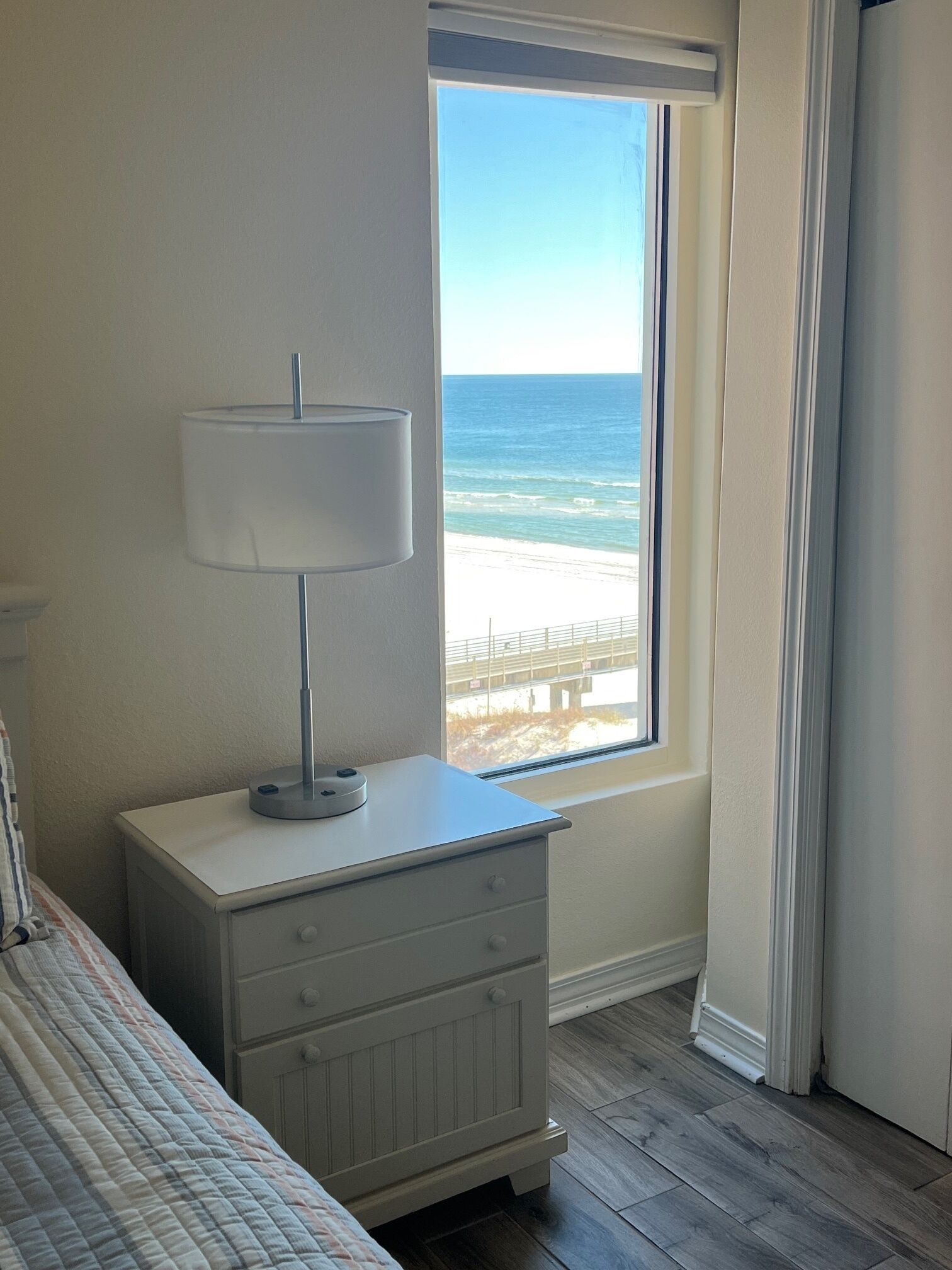 A bedroom with a bed, a bedside table with a lamp, a window with a view of the beach and ocean.