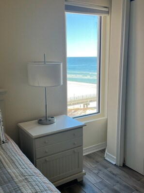 Quarto com cama, mesa de cabeceira com luminária, janela com vista para a praia e oceano.