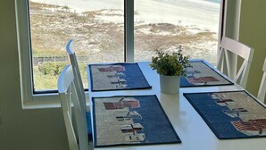 A dining table with blue placemats featuring American flags, white chairs, and a view of a beach through a large window. - Save 10% Last min & Week! Gulf Front, Panoramic Views, Beach Supplies, Pool! (Orange Beach)