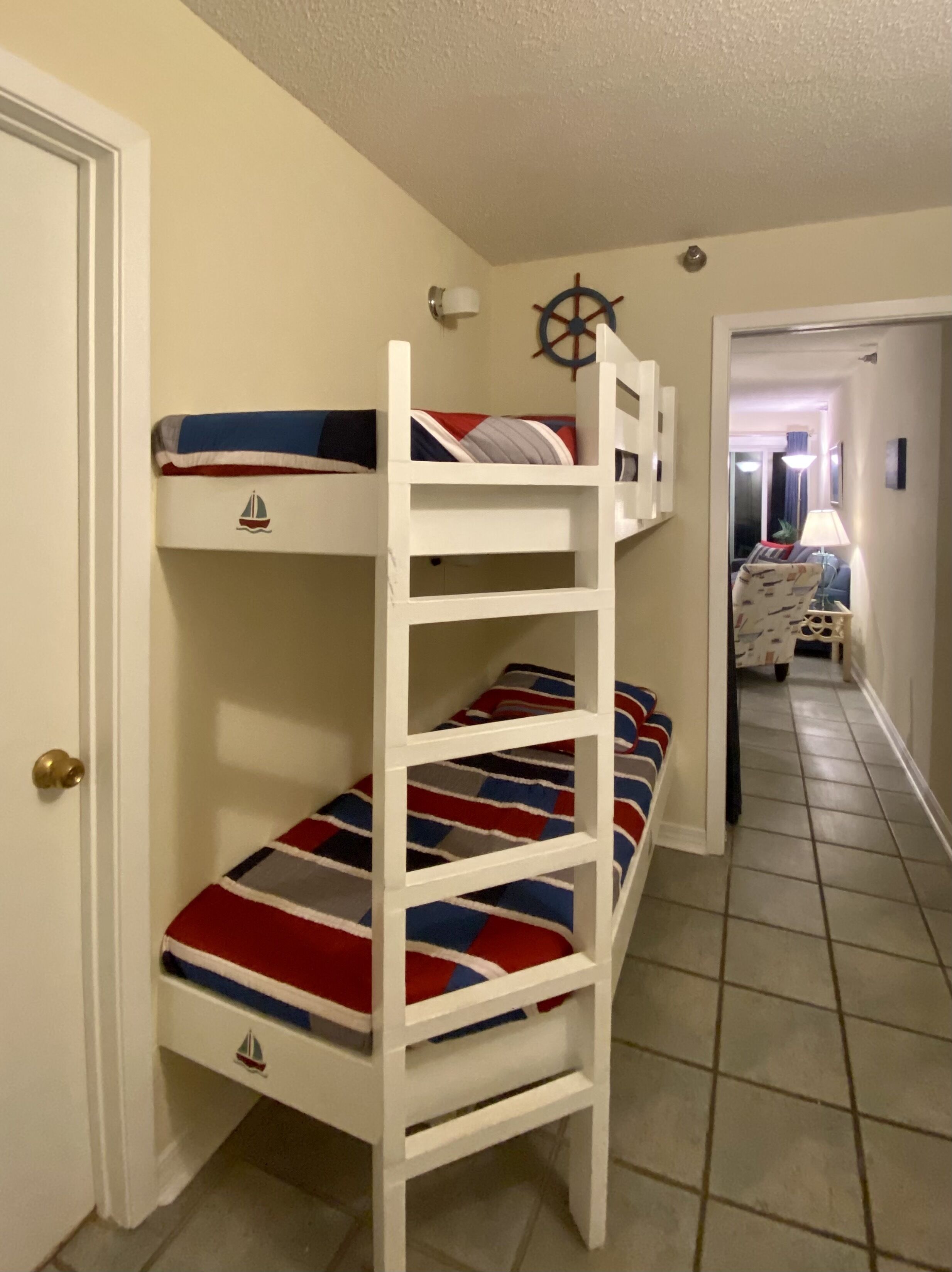 A bunk bed with a striped bedspread, a wall-mounted ship's wheel, and a door with a brass doorknob.