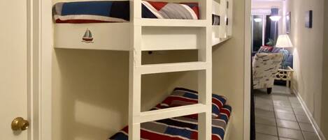 A bunk bed with a striped bedspread, a wall-mounted ship's wheel, and a door with a brass doorknob.