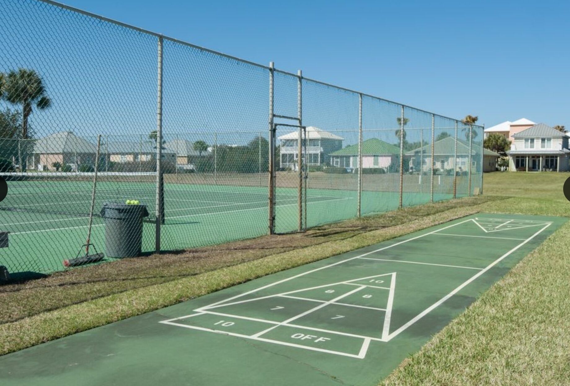 Un court de tennis entouré d’une clôture en treillis métallique, avec un terrain de jeu de shuffleboard situé à côté.