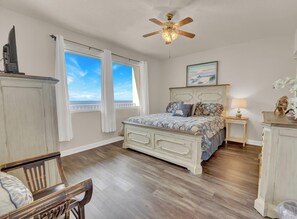 A bedroom with a bed, a ceiling fan, a window with curtains, a nightstand with a lamp, and a painting on the wall.