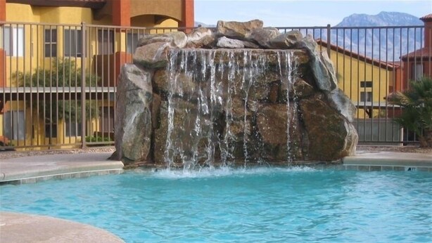 A swimming pool with a waterfall feature, surrounded by a fence and buildings in the background. - Clean & Cozy Condo with pool and trailer parking! (Mesquite)