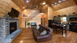 Un interior acogedor de cabaña con chimenea de piedra, sofá de cuero, mesa de comedor, zona de cocina y escalera de madera.