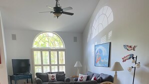 A living room with a gray sofa, wooden dining table, and chairs. There is a television on a stand, a lamp, and a window with a view of trees. - Updated with everything you need for family fun time, just minutes to beach! (Ocean View)