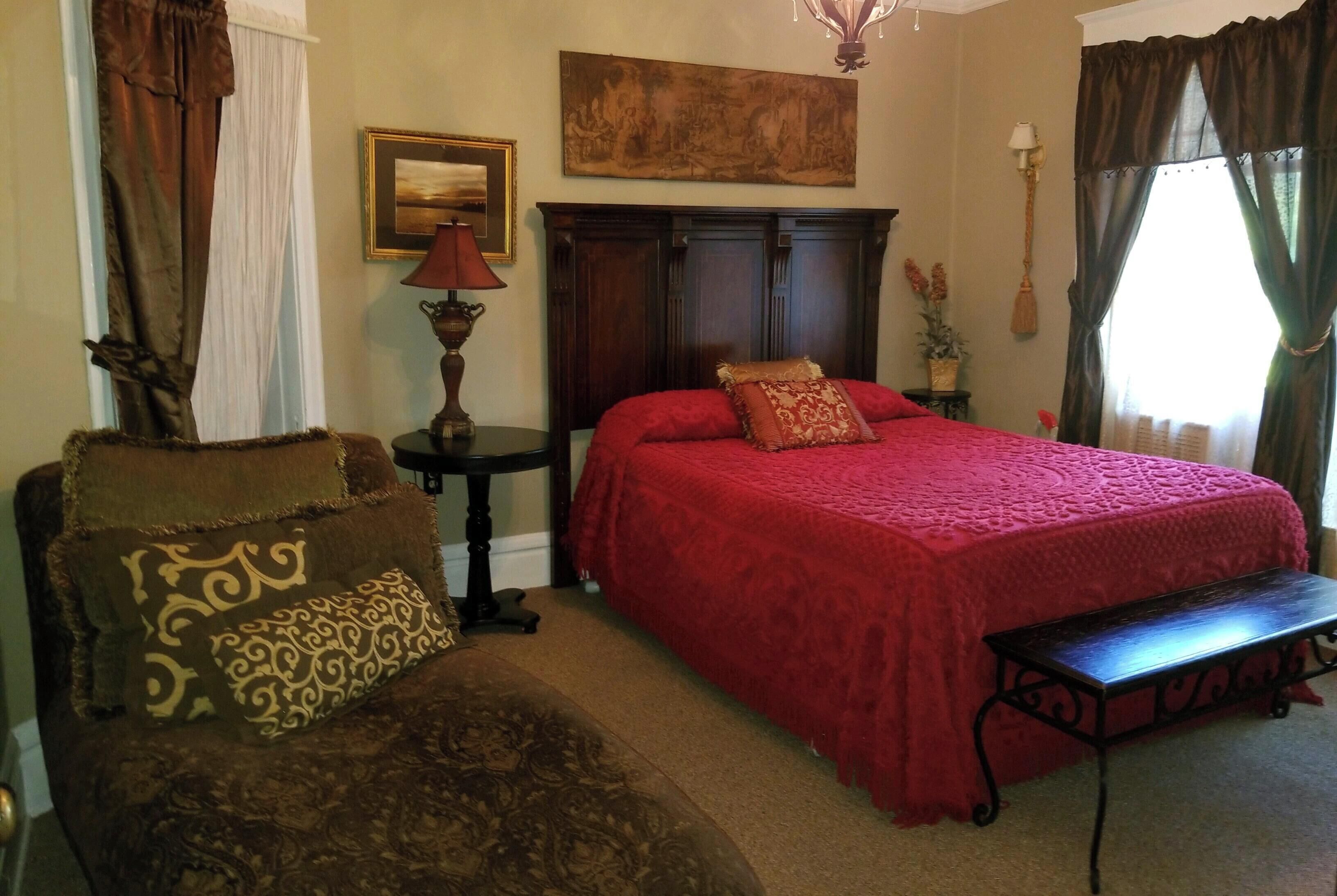 A bedroom with a large bed, a wooden nightstand, a chair, a bench, a chandelier, a painting, and a window with curtains.