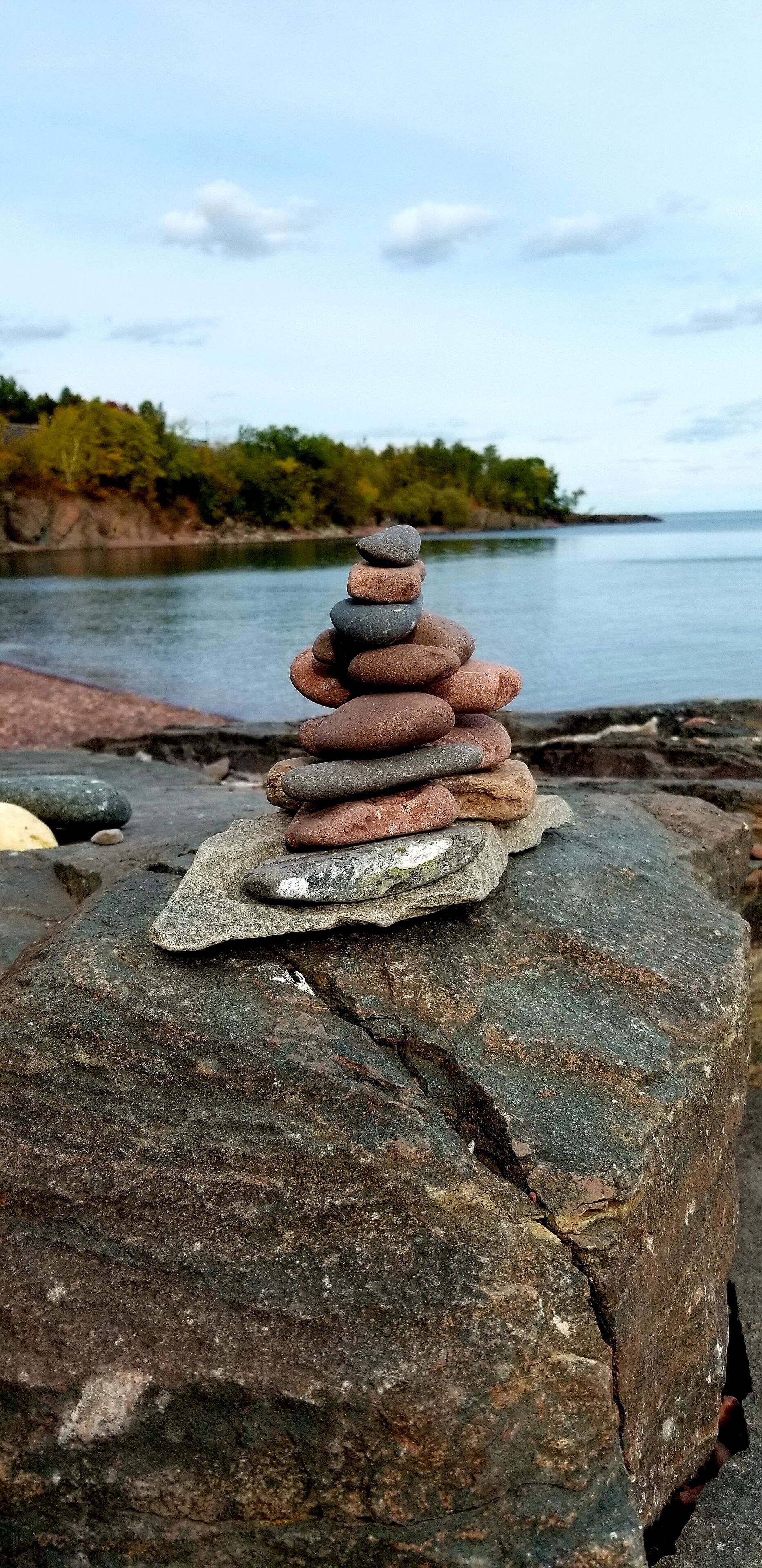 Une pile de pierres polies, empilées avec équilibre sur un grand rocher, au bord d’une étendue d’eau calme, avec une rive boisée en arrière-plan.