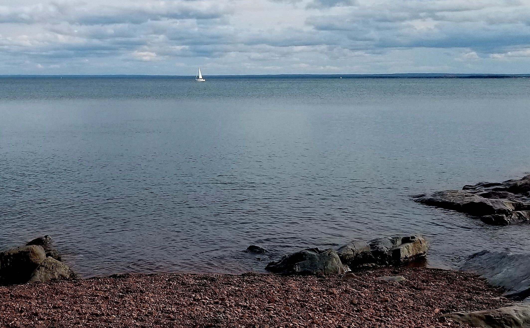 Ruhige See mit einem Segelboot in der Ferne, felsige Küstenlinie und bewölkter Himmel.