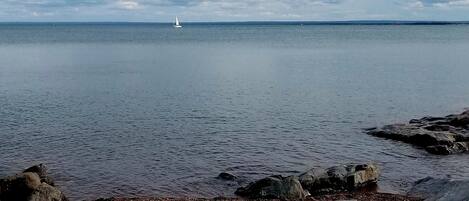 Ruhige See mit einem Segelboot in der Ferne, felsige Küstenlinie und bewölkter Himmel.
