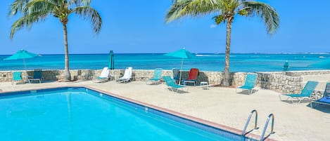 A swimming pool with lounge chairs and umbrellas, palm trees, and a view of the ocean.
