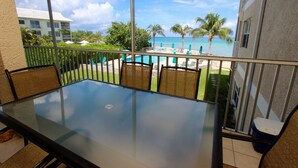 Ein Balkon mit Glastisch und Stühlen mit Blick auf einen Pool und Strand, Palmen und ein Gebäude im Hintergrund.