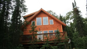 A two-story wooden cabin with a balcony, surrounded by dense forest.