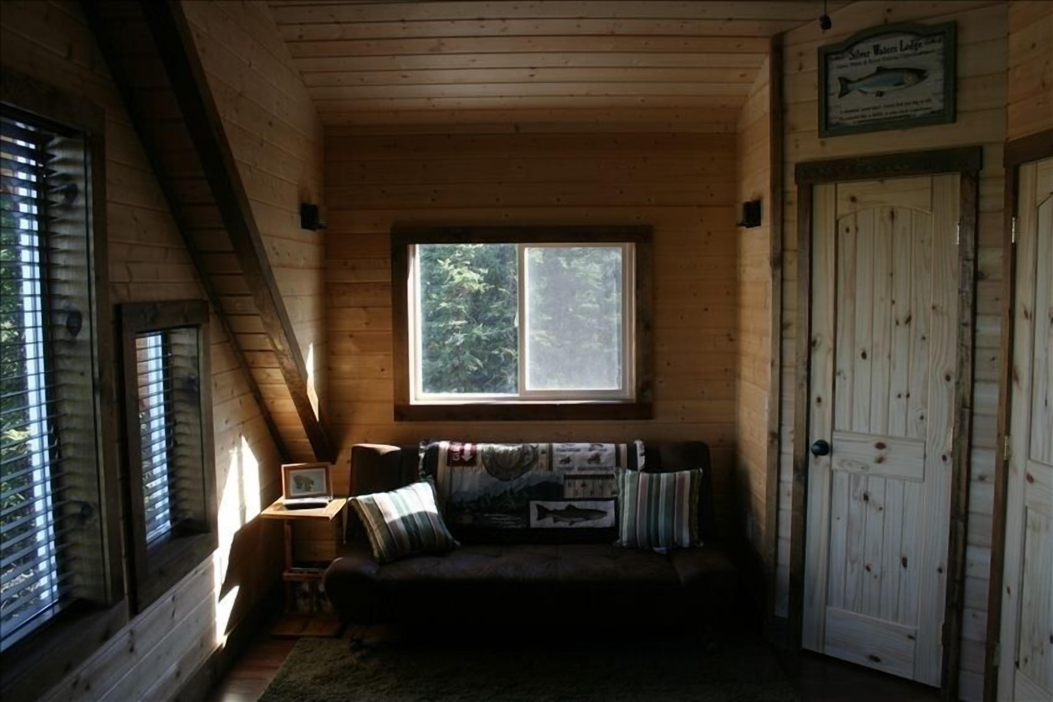 Interior acollidor d'una cabana amb sostre de fusta, una finestra amb vista a arbres, un sofà amb almoadins i una porta amb un senyal de peix.