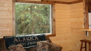 A cozy cabin interior with a wooden interior, a large window with a view of trees, a couch with a 'Alaska' themed pillow, and a wooden staircase.
