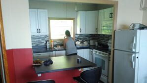 A compact kitchen with white cabinets, a black countertop, and a refrigerator. There are two black bar stools and a window providing natural light. - MacEachern 19th Hole Celtic House (Creignish)