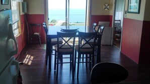 A dining area with a table and chairs, a view of the sea through a large window, and a clock on the wall. - MacEachern 19th Hole Celtic House (Creignish)