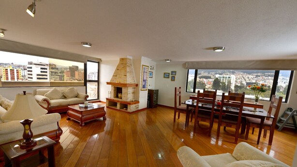 A spacious living room with wooden flooring, a fireplace, a dining area with wooden chairs, and large windows offering city views. - Exc PH, Best Loc, Near Carolina, Huge Balcony, AMZ Views, WiFi, APU, 💲Deals! (Quito Canton)