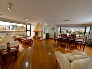 A spacious living room with wooden flooring, a fireplace, a dining area with wooden chairs, and large windows offering city views. - Exc PH, Best Loc, Near Carolina, Huge Balcony, AMZ Views, WiFi, APU, 💲Deals! (Quito Canton)