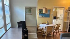 Una habitación con un escritorio de madera, una mesa de comedor con sillas de mimbre, un sofá y una cocina visible a través de una puerta abierta.