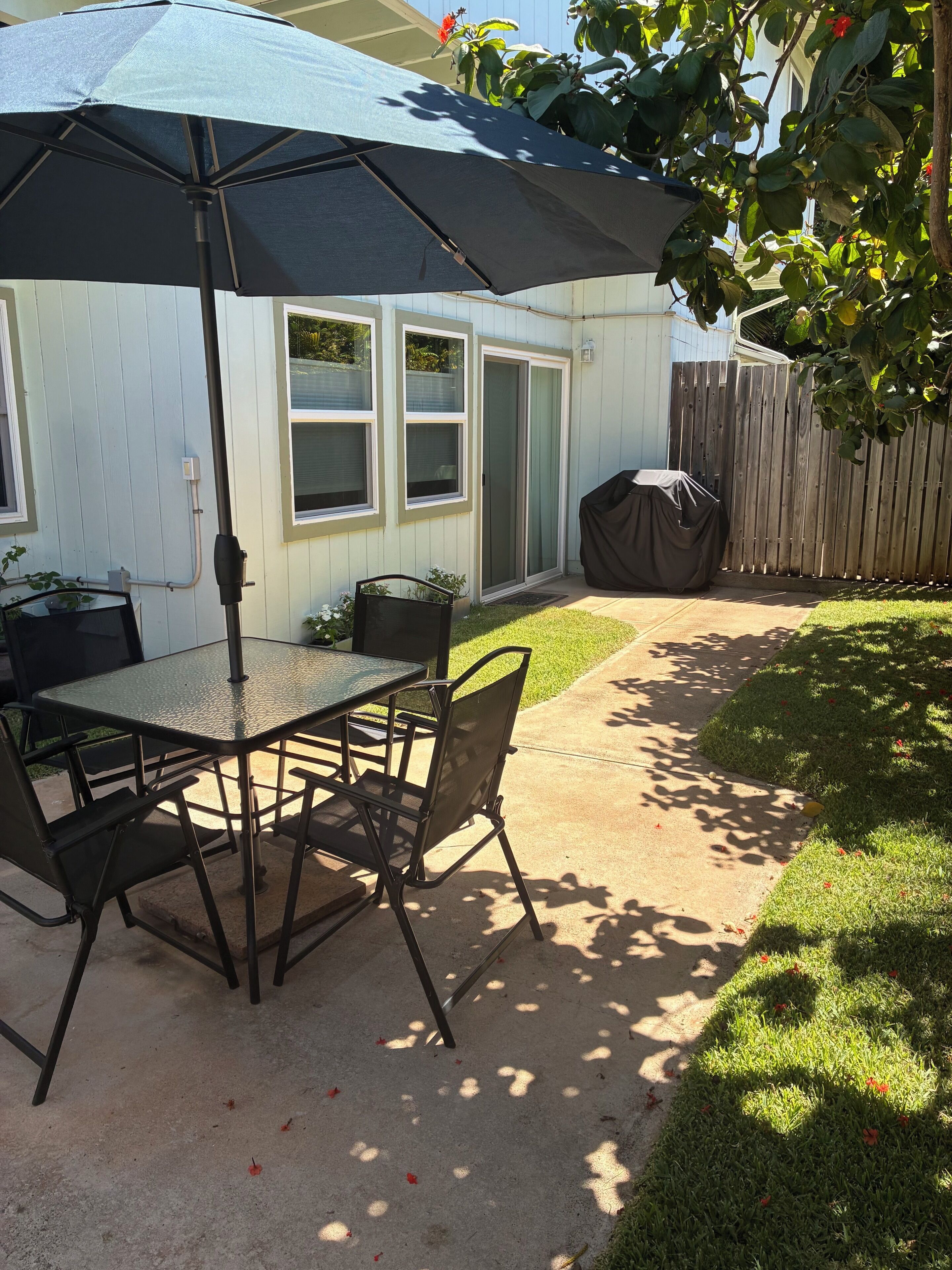 An outdoor patio with a table and chairs, a covered grill, and a wooden fence.