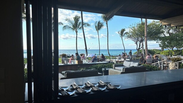 A view from a covered outdoor area with tables and chairs, overlooking a beach with palm trees and the ocean. - Enjoy a Hawaiian vacation in Paradise Stay @ The World Famous Mauna Lani Resort (Puako)