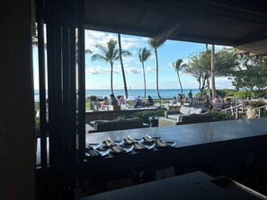 A view from a covered outdoor area with tables and chairs, overlooking a beach with palm trees and the ocean. - Enjoy a Hawaiian vacation in Paradise Stay @ The World Famous Mauna Lani Resort (Puako)