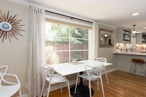 Une cuisine moderne avec une table et des chaises blanches, une grande fenêtre avec des rideaux et une horloge murale au motif rayonnant.