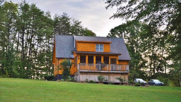 Une maison en bois à deux étages, dotée d’un balcon, entourée d’une cour verdoyante et d’arbres.