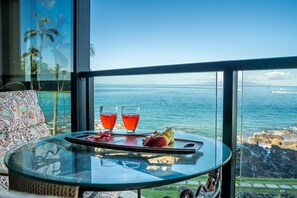 A glass table with a tray holding two glasses of red beverage, a fruit, and a small dish, set against a scenic ocean view. - 202 - Premium remodeled oceanfront with A/C - Spectacular! (Kihei)