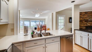 A modern kitchen with white cabinetry, a stainless steel sink, and a dishwasher. There is a brick wall with a fireplace and a large window overlooking a lake. - BREATHTAKING VIEWS AT NORMANDY BREEZE! (Denver)