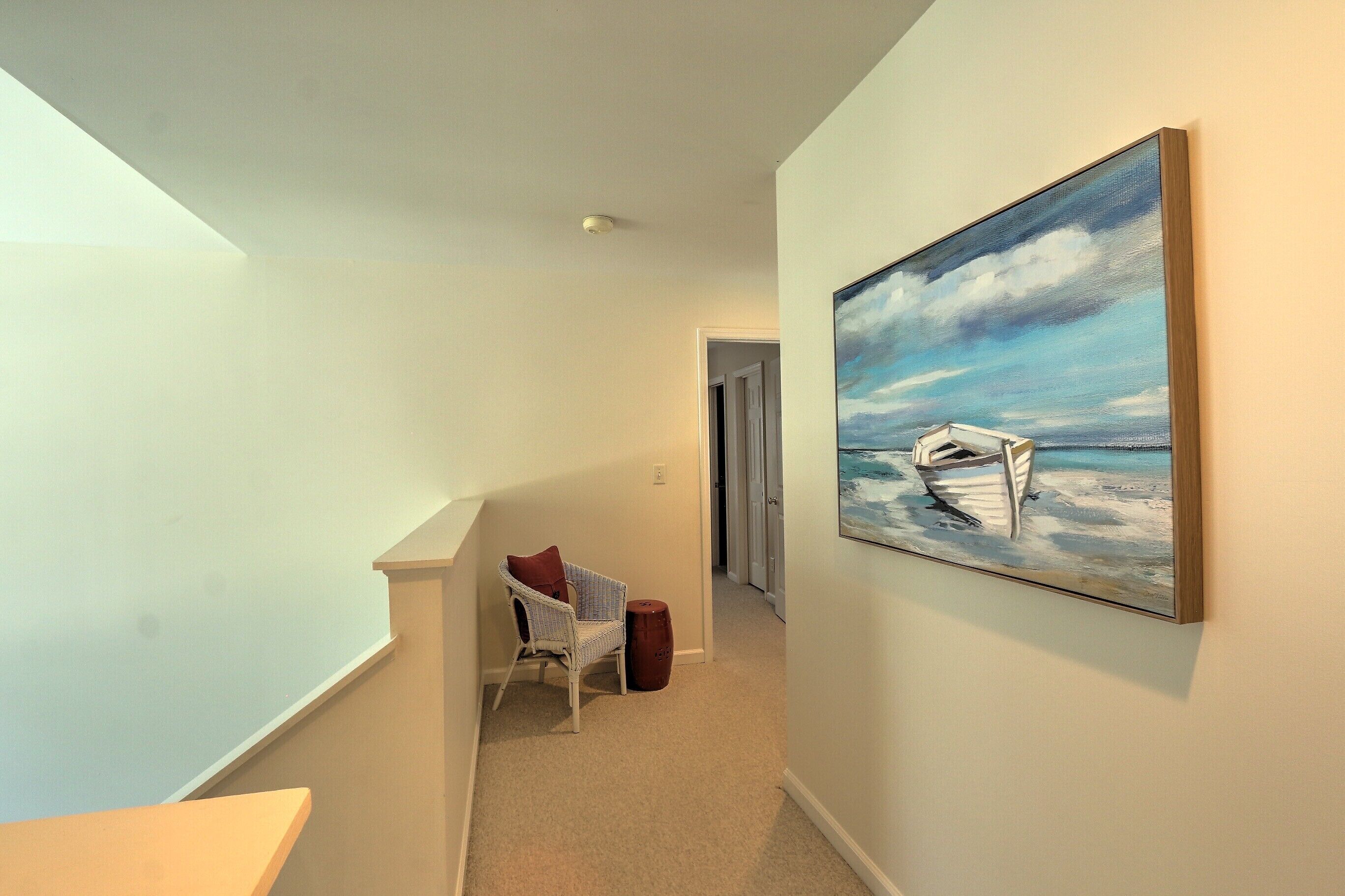 A hallway with a painting of a boat on a beach, a chair with a cushion, and a small wooden stool.