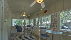 A sunlit porch with white wooden walls, a ceiling fan, and a large window offering a view of the outdoors.