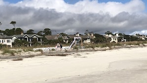 A beach with a clear sky, houses in the background, and a sandy area with some vegetation.