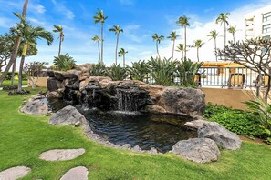A landscaped garden with a small waterfall, palm trees, and a building in the background. - Squeaky clean ground floor Sugar Beach #104 (Kihei)