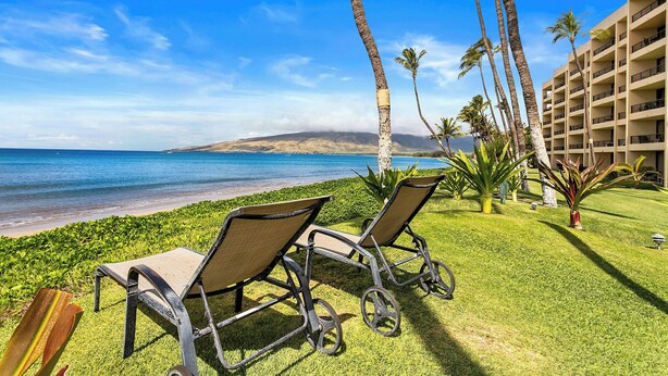 A beach view with lounge chairs, palm trees, and a building in the background. - Squeaky clean groundfloor Sugar Beach #104 (Kihei)
