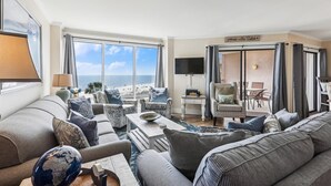 Ein geräumiges Wohnzimmer mit großem Fenster mit Blick auf einen Strand, ausgestattet mit einer Couch, Sesseln und einem Couchtisch.