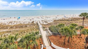 Ein Strand mit einer hölzernen Promenade, die zum Ufer führt, umgeben von Dünen und Palmen.