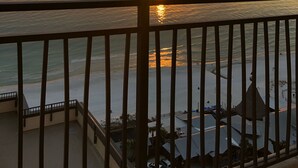 A balcony with a view of the sunset over the ocean, a railing, and a building with a thatched roof in the foreground.