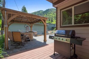 Eine Terrasse im Freien mit Grill, einer überdachten Sitzgruppe und Blick auf einen bewaldeten Hügel.