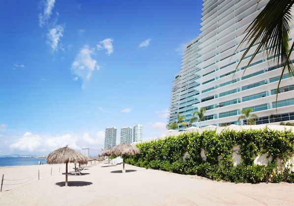 Un hotel frente a la playa con una palmera, un paraguas de paja y vista al océano.