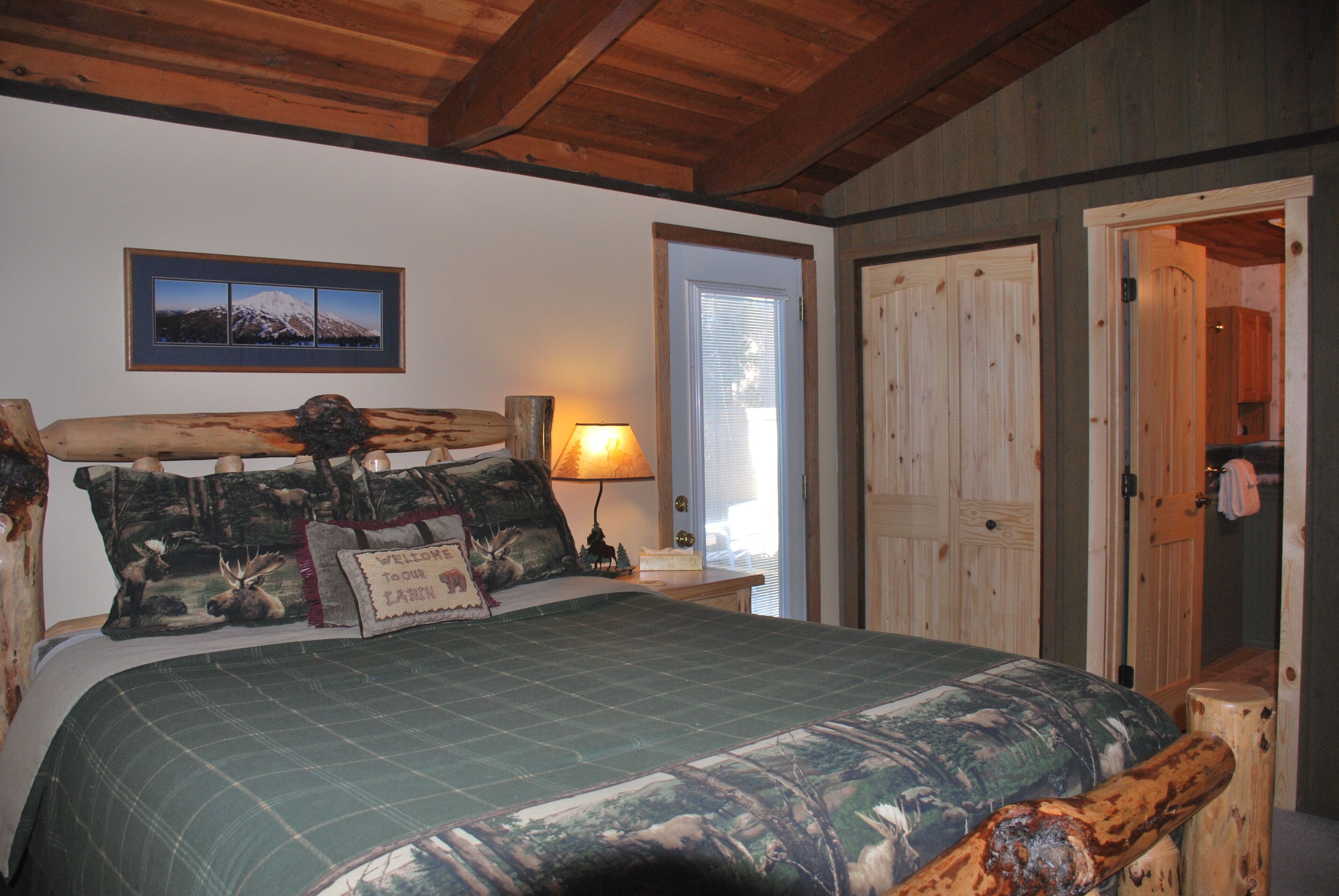 Ein Schlafzimmer mit einem hölzernen Kopfende, einem Bett mit Bettdecke im Tarnmuster, einem gerahmten Bild an der Wand, einer Nachttischlampe und einer Tür zu einem anderen Raum.