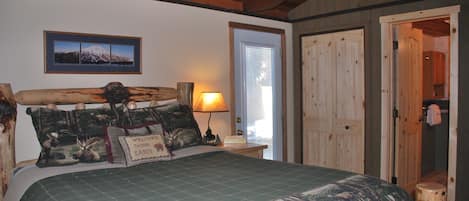Ein Schlafzimmer mit einem hölzernen Kopfende, einem Bett mit Bettdecke im Tarnmuster, einem gerahmten Bild an der Wand, einer Nachttischlampe und einer Tür zu einem anderen Raum.