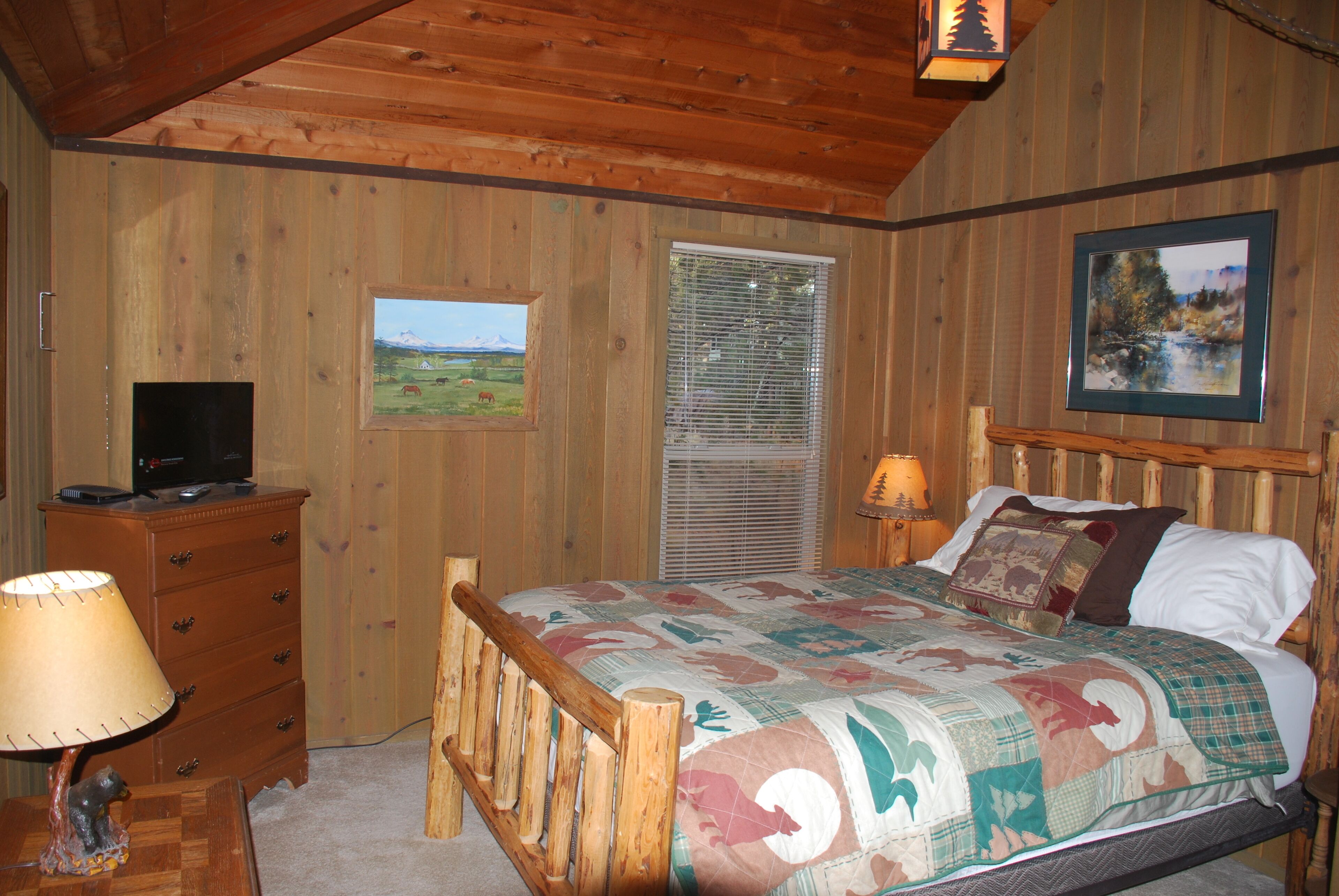 Un dormitorio con una cama de madera, un aparador, un televisor sobre el aparador y una ventana con persianas.