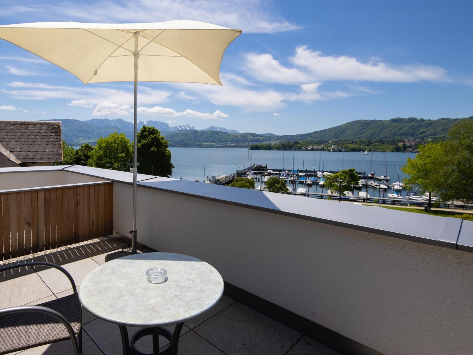 Ein Balkon mit Tisch und Stuhl, einem großen Sonnenschirm und Blick auf einen Jachthafen und Berge.