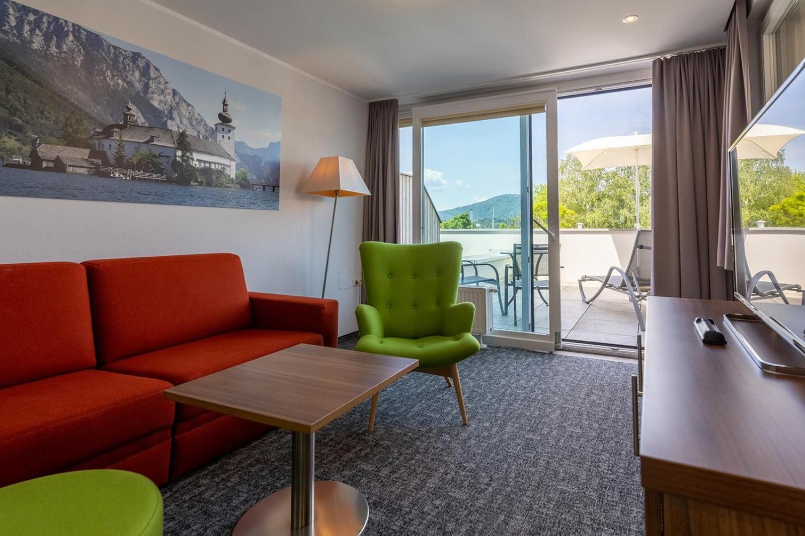 A hotel room with a red sofa, a green armchair, a small table, a lamp, and a view of the outdoors through a sliding glass door.