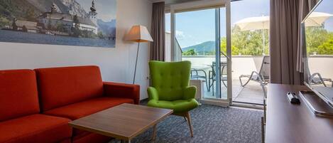 A hotel room with a red sofa, a green armchair, a small table, a lamp, and a view of the outdoors through a sliding glass door.