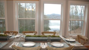 Un coin repas avec une table dressée pour quatre personnes, de grandes fenêtres offrant une vue sur un lac et une forêt, et un pot de fleurs avec des plantes vertes placé sur la table.