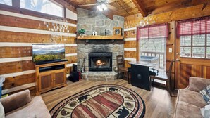 Un interior de cabaña acogedor, con chimenea de piedra, paredes de madera, televisor y un tapete redondo con motivos de animales.